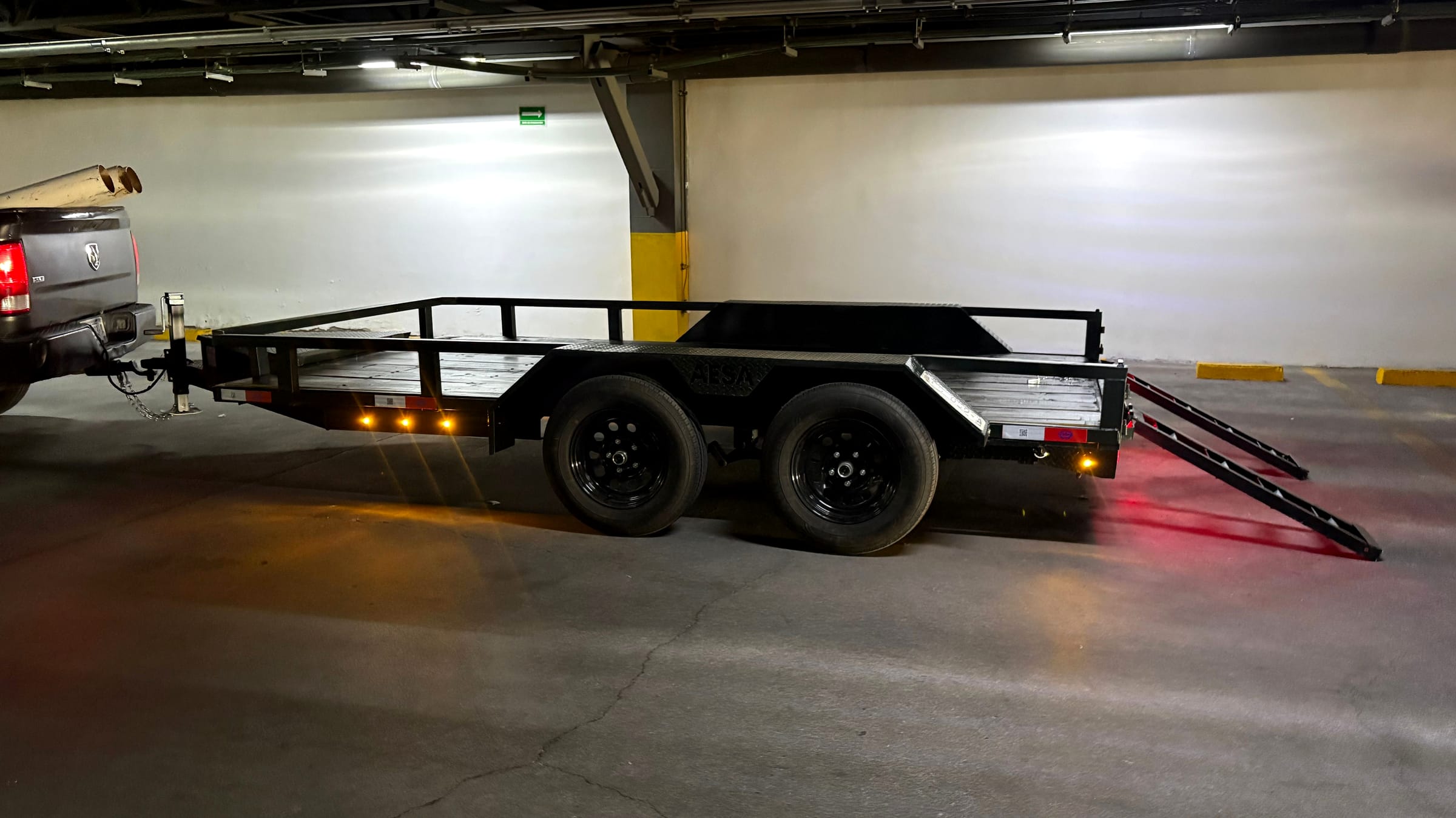 Remolque de plataforma negro con barandal y luces LED encendidas en un estacionamiento techado